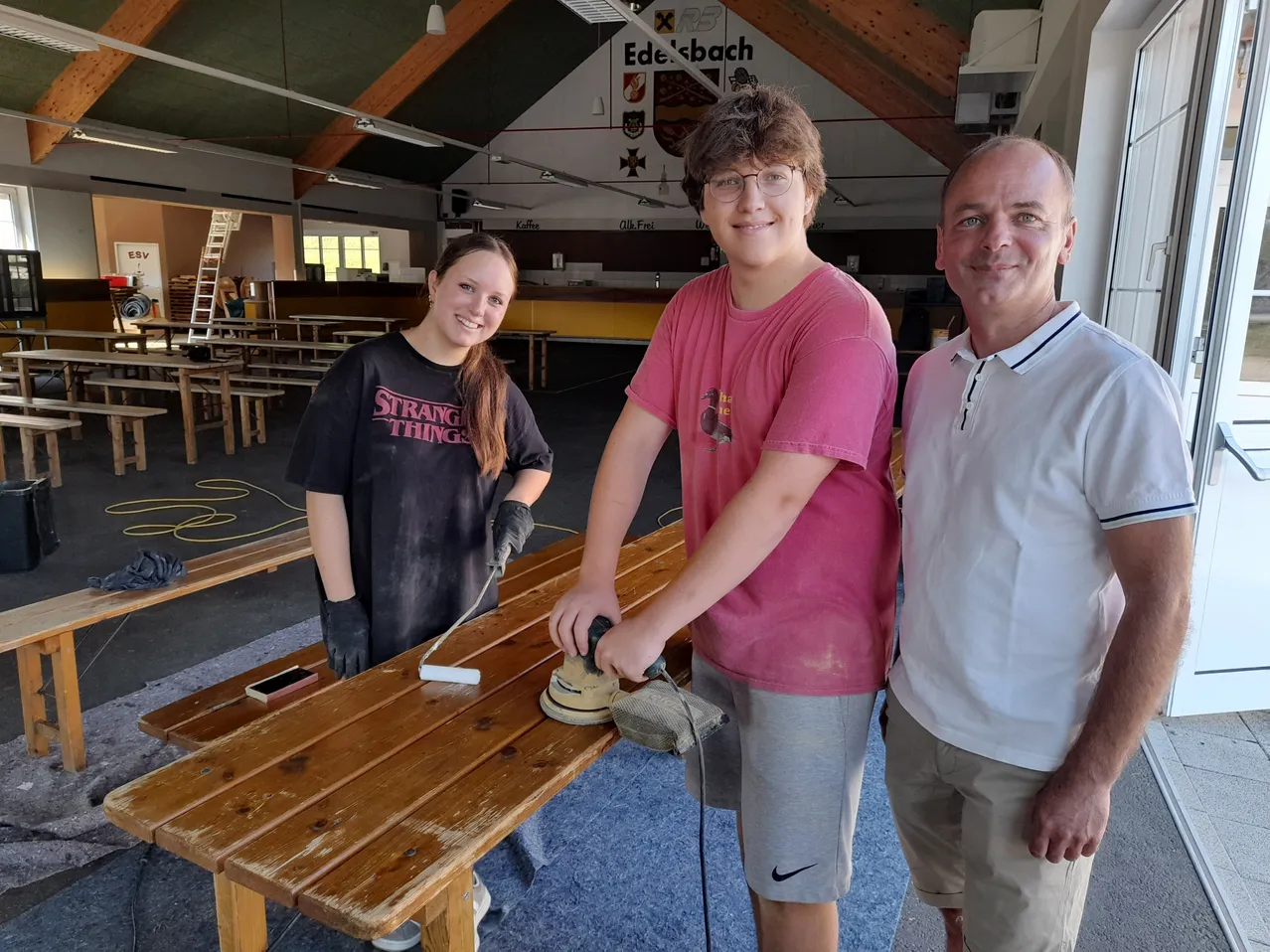 Drei Personen stehen um einen Holztisch, zwei von ihnen lächeln und einer benutzt einen Schleifer auf dem Tisch.