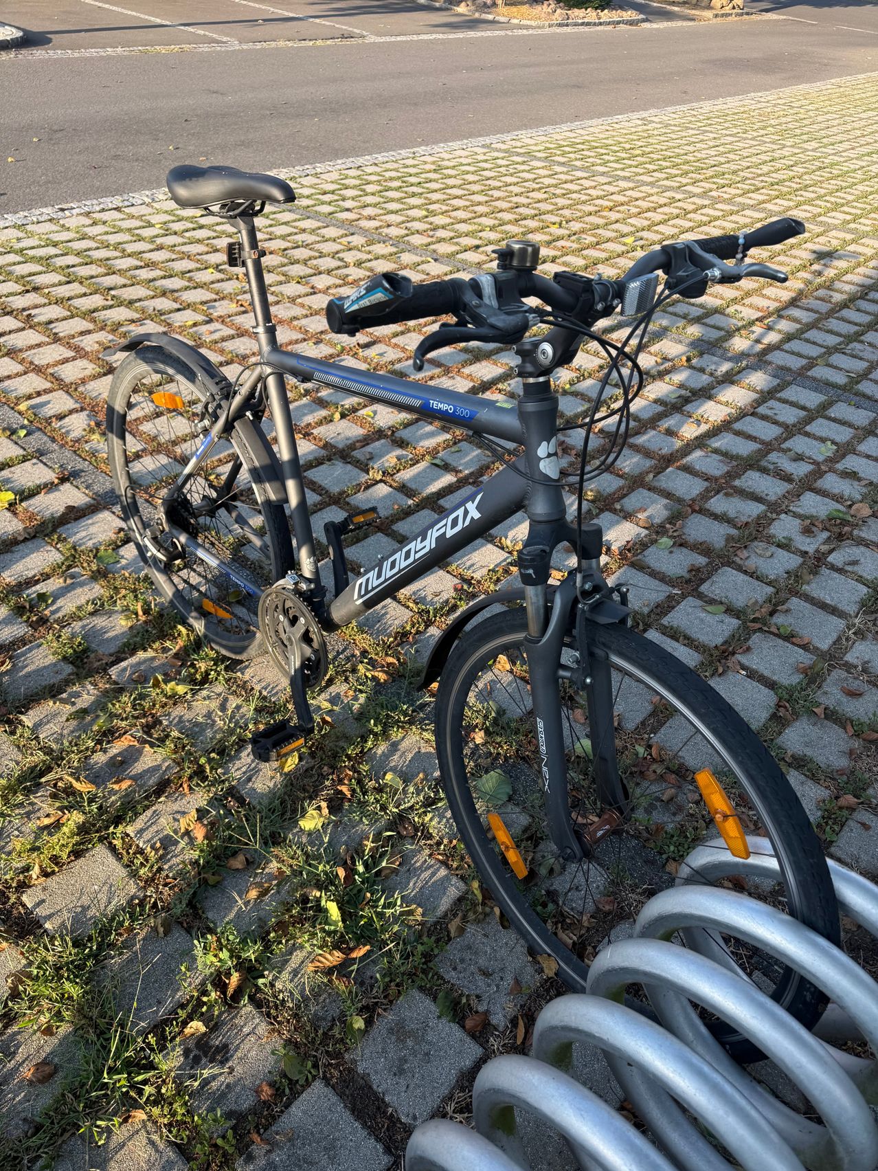 A black Mudgyfox Tempo 300 bicycle is parked on a brick pathway with yellow reflectors on the wheels.