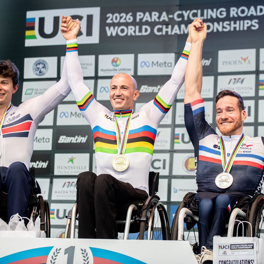 Drei Rollstuhlfahrer auf dem Podium feiern, tragen Radtrikots und Medaillen. Die zentrale Figur hat eine goldene Medaille um den Hals. Hinter ihnen ist ein Hintergrund mit dem Logo der Para-Cycling Road World Championships 2026.