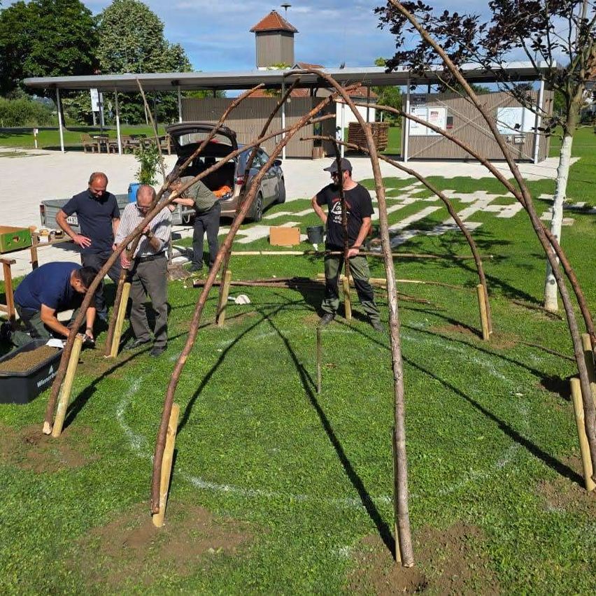 Five men work on a wooden structure in a grassy area. One man bends over, another holds a tool, while the others stand. A car is parked nearby, and trees are in the background.