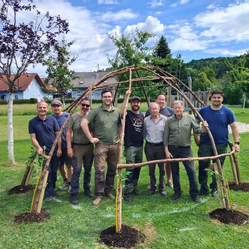 A group of men pose with a small, newly constructed wooden shelter in a park, with houses and trees in the background.