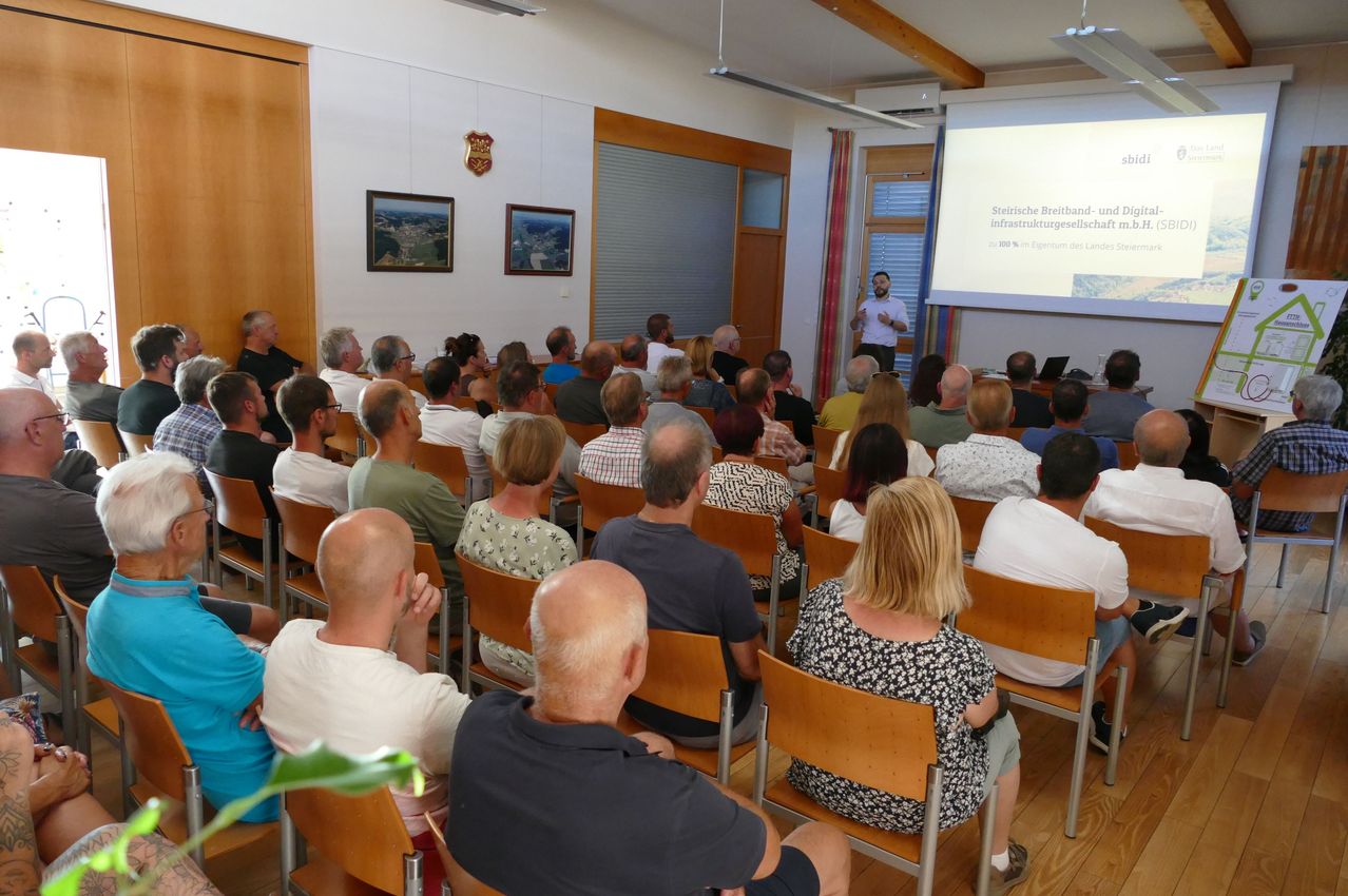 A presentation is taking place in a room with a projector screen. A man is standing in front of a projector screen, while the audience is seated in rows.