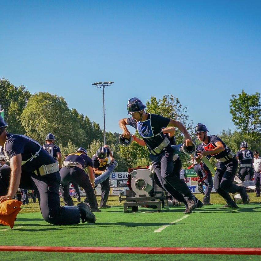 A group of individuals are participating in a team sport on a green field. They wear helmets and uniforms, and one is pulling a wheel with a rope. The setting includes a red line and trees in the background.