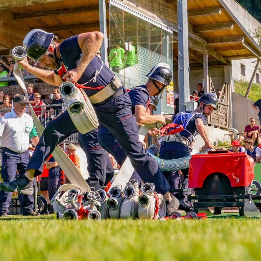 Firefighters in protective gear are practicing their skills in an outdoor setting with a crowd watching. One man is holding a hose while another is connected to a fire engine.