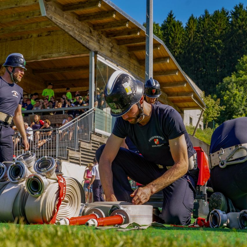 A group of firefighters is preparing equipment for a demonstration in front of a building. A crowd is watching from the bleachers.