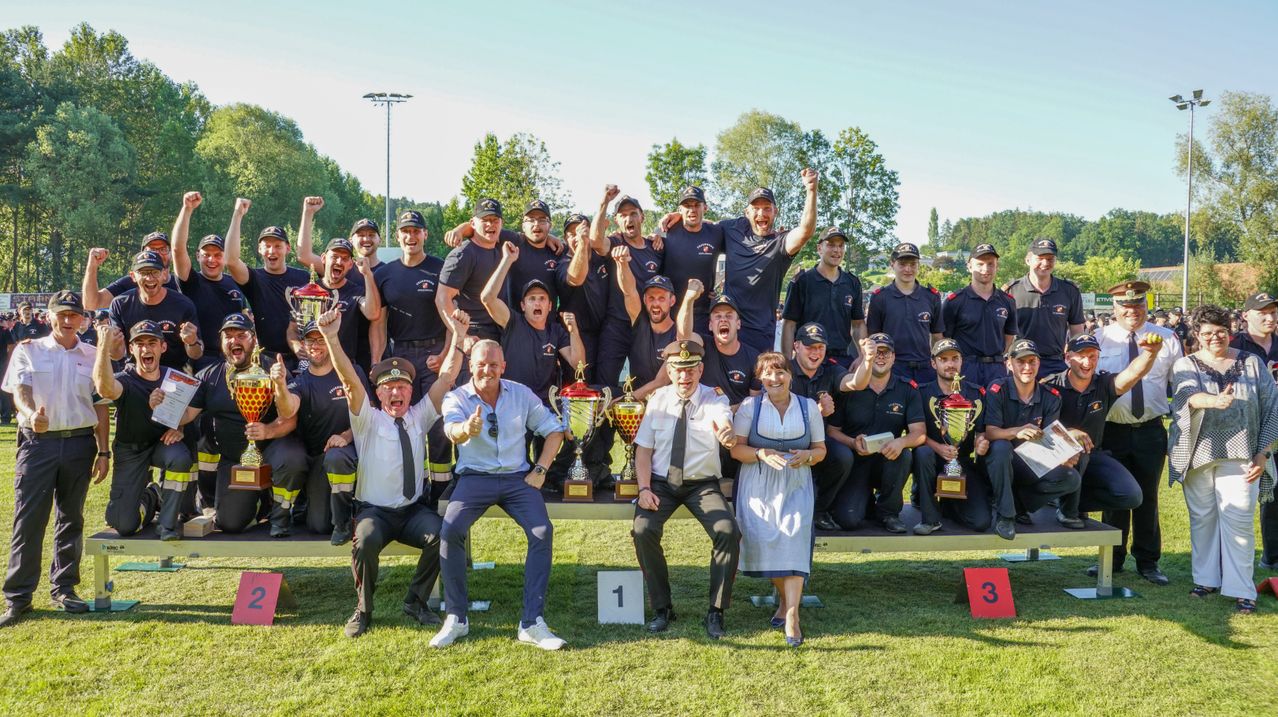 A group of firefighters celebrates with trophies in a sunny outdoor setting. They pose with joy and pride, surrounded by trees and a grassy field.