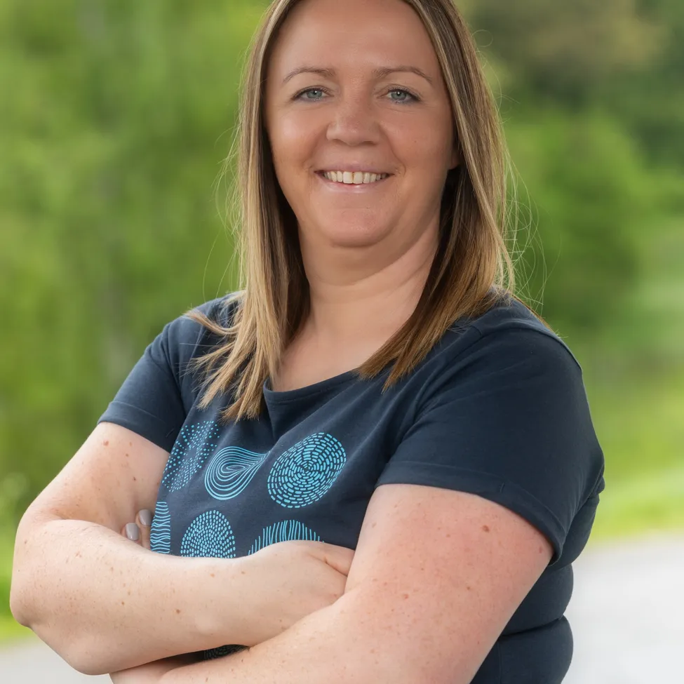 Eine lächelnde Frau mit blonden Haaren, die ein blaues T-Shirt mit einem kreisförmigen Design trägt, steht mit verschränkten Armen in einer grünen Außenumgebung.