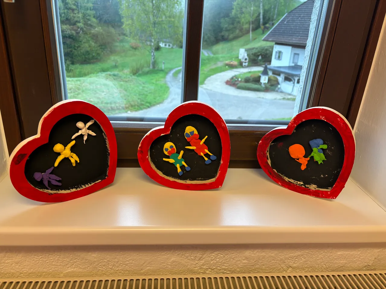 Drei herzförmige Bastelarbeiten mit bunten Figuren stehen auf einer Fensterbank. Dahinter ist ein Fenster mit Blick auf eine Wiese und ein Haus.
