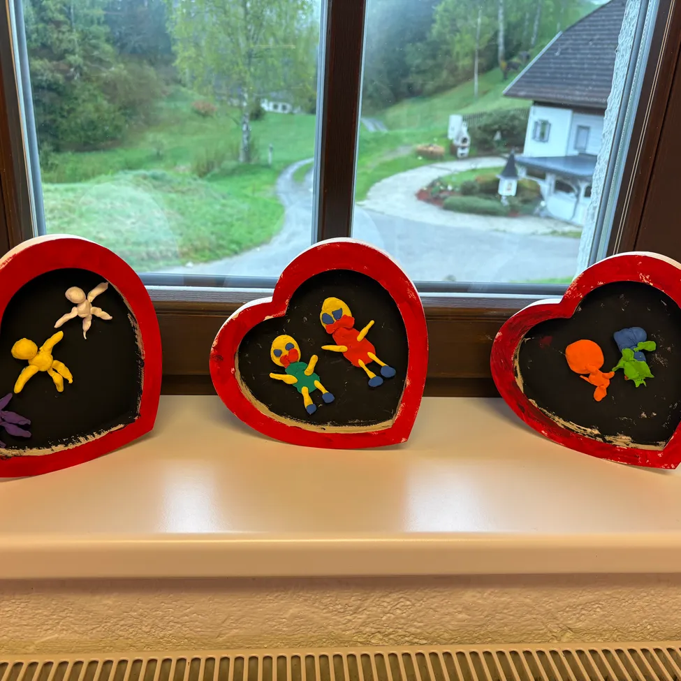 Drei herzförmige Bastelarbeiten mit bunten Figuren stehen auf einer Fensterbank. Dahinter ist ein Fenster mit Blick auf eine Wiese und ein Haus.