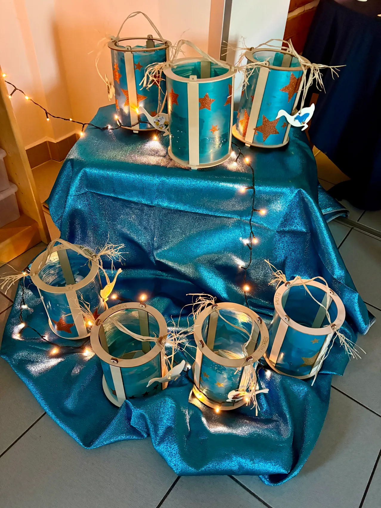 Ein Tisch, der mit einem blauen Tuch bedeckt ist, hat mehrere beleuchtete blaue Laternen mit Sternen und Schwänen. Der Hintergrund ist ein Holzregal mit einer Aufbewahrungsbox.