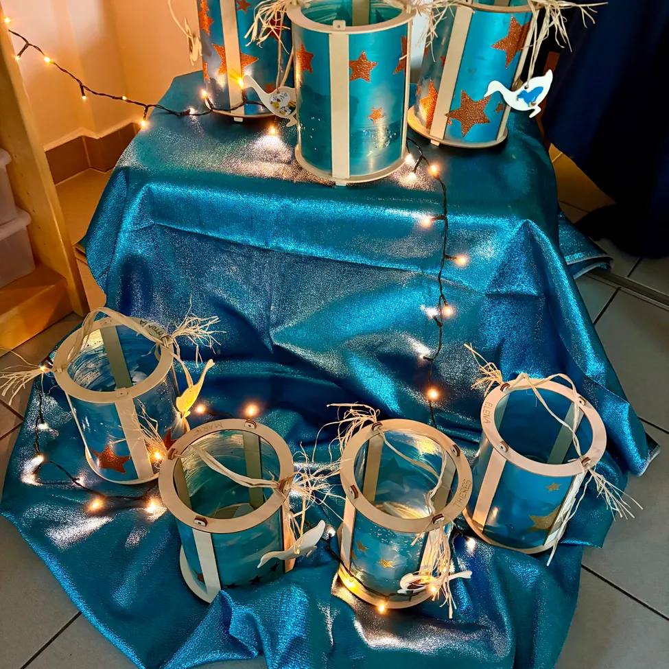 Ein Tisch, der mit einem blauen Tuch bedeckt ist, hat mehrere beleuchtete blaue Laternen mit Sternen und Schwänen. Der Hintergrund ist ein Holzregal mit einer Aufbewahrungsbox.