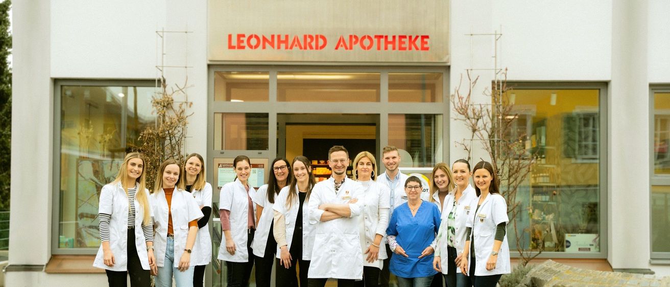 Eine Gruppe von Menschen in Laborkitteln posiert vor einem Apothekengebäude. Das Gebäude heißt Leonhard Apotheke.