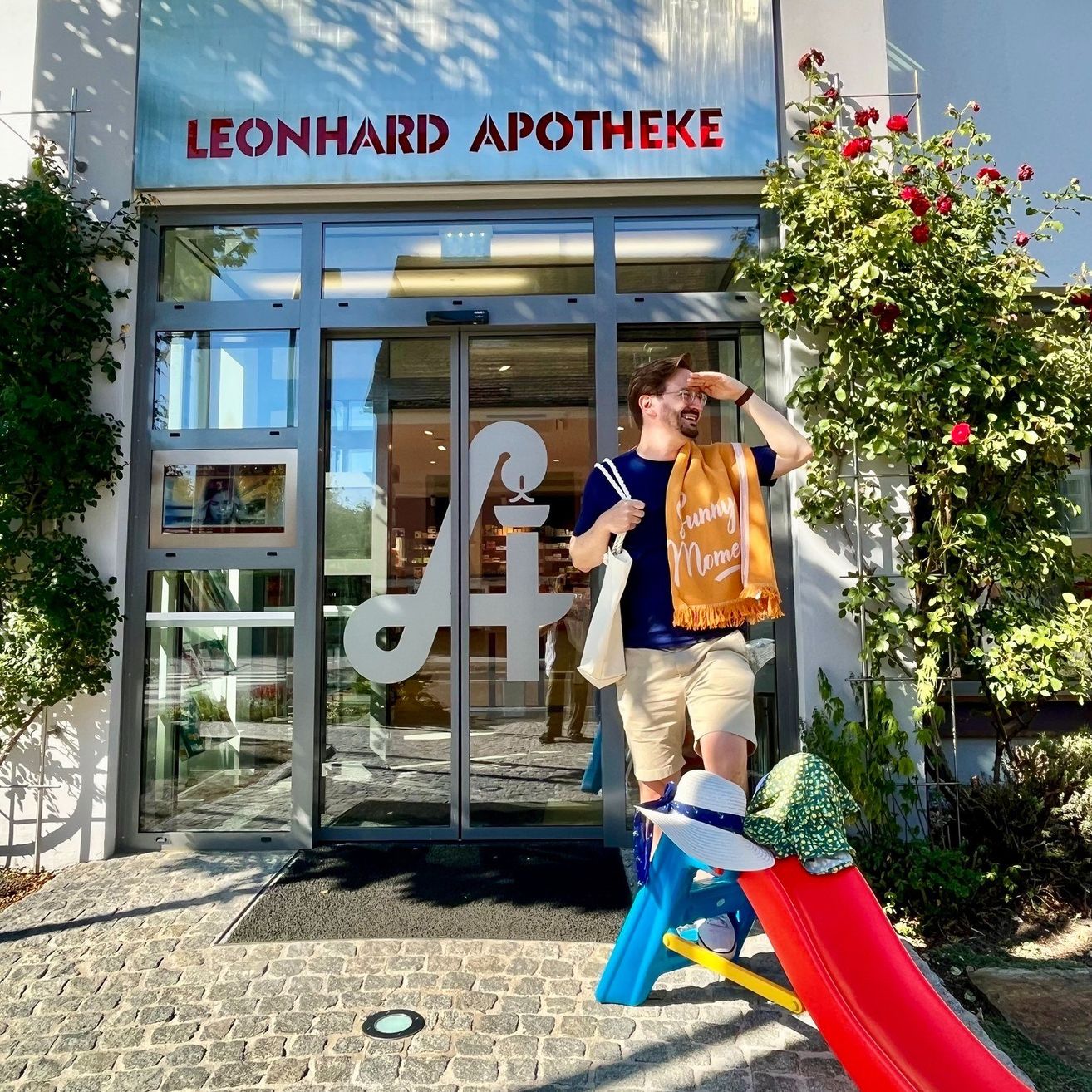 Ein Mann steht vor der Leonhard Apotheke, lächelt und posiert für ein Foto. Er hält eine Tasche und trägt einen Schal, mit einem bunten Schlitten hinter ihm.