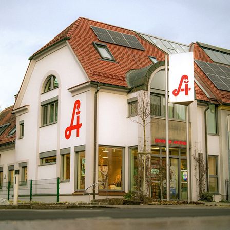 Ein weißes Gebäude mit rotem Dach, Solarpanelen und einem roten Buchstaben 'A' an der Seite. Es hat Glasfenster und eine Glastür mit einem Schild, das 'Leonhard Apotheke' steht.