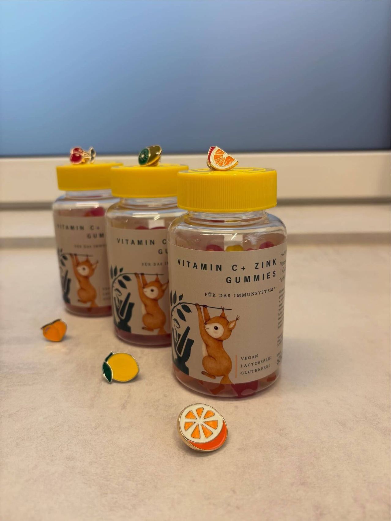 Drei Flaschen Vitamin C und Zink-Gummis stehen auf einem Tisch. Jede Flasche hat einen gelben Deckel mit einer fruchtthematischen Anstecknadel obenauf.