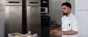 Ein Koch steht in einer Küche mit zwei Edelstahlkühlschränken und erklärt etwas mit einem Tisch voller verschiedener Zutaten vor ihm.