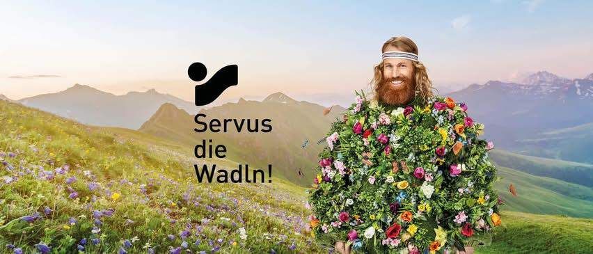 Ein Mann mit Bart steht in einem Blumenfeld. Hinter ihm steht ein Logo, das 'Servus die Wadln!' sagt. Im Hintergrund sind Berge.