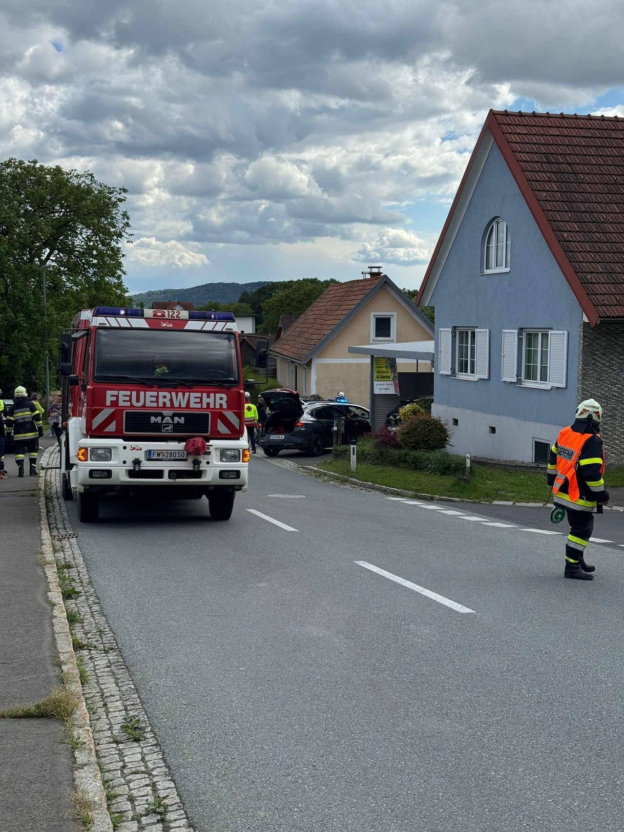 Ein Feuerwehrwagen ist am Straßenrand geparkt, mit Feuerwehrleuten und einer Person in einer orangefarbenen Weste in der Nähe. Die Szene umfasst auch ein demoliertes Auto und Wohngebäude.