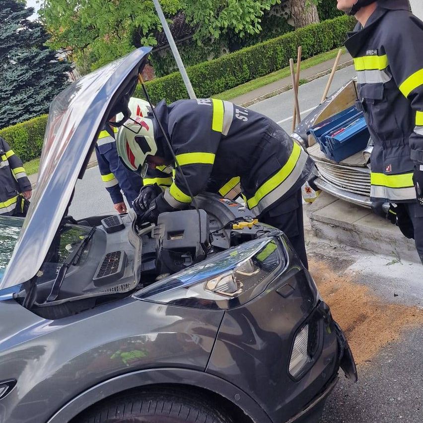 Feuerwehrleute inspizieren ein beschädigtes Auto mit offenem Motorhaube am Straßenrand. Ein Werkzeugkasten befindet sich in der Nähe, und ein kleiner Haufen Kies liegt auf dem Boden.
