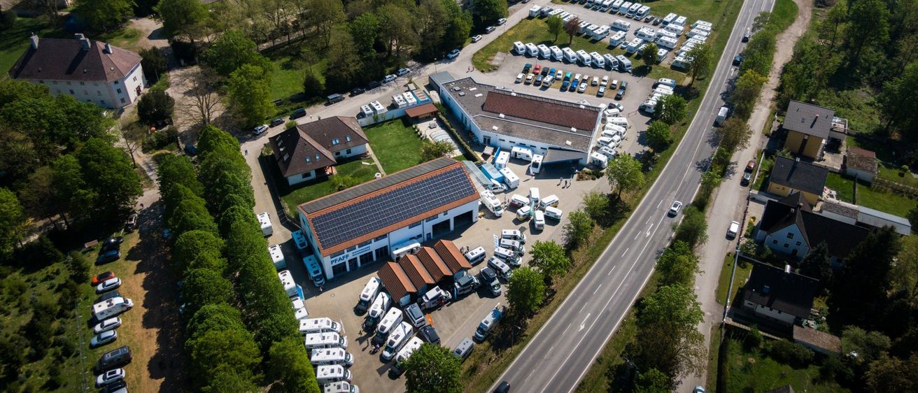 Luftaufnahme eines Campingplatzes mit mehreren geparkten Wohnmobilen, Solarpanelen auf dem Dach, einer Straße mit Autos und einem Ackerfeld im Hintergrund.