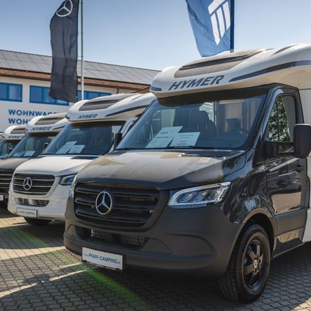 Eine Reihe von Hymer Mercedes-Benz Wohnmobilen, die vor einem Gebäude geparkt sind. Der vordere Van ist schwarz mit weißen Akzenten, während die anderen weiß sind.