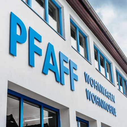 Ein weißer Bau mit blauer Einfassung trägt in großen blauen Buchstaben 'PFAFF'. Das Gebäude hat mehrere Fenster und eine blau umrahmte Eingangstür. Unter dem Schild steht 'WOHNWAGENWOHNMOBILE.'