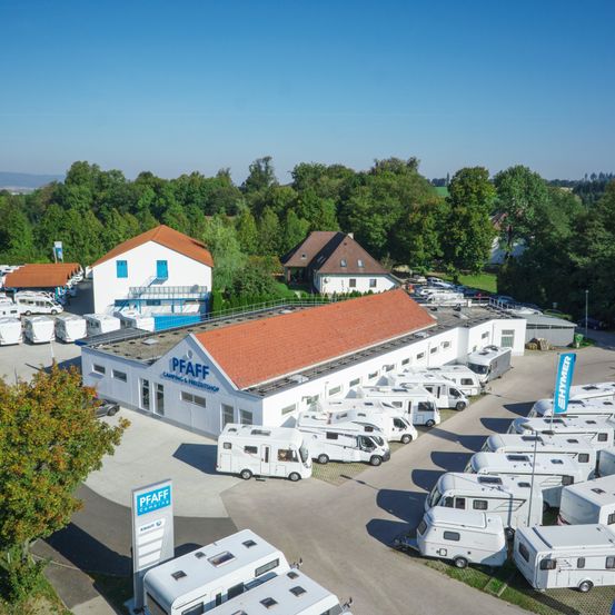 Ein Luftbild eines weissen Gebäudes mit rotem Dach, beschriftet mit 'PFAFF CAMPING & FAHRZEUGSHOP'. Viele Wohnmobile sind davor geparkt.