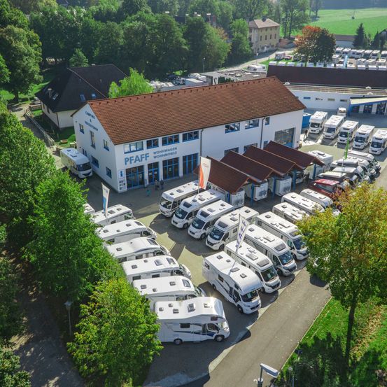 Eine Luftaufnahme eines Parkplatzes mit vielen weißen Wohnmobilen. In der Mitte befindet sich ein Gebäude mit der Aufschrift PFAFF Wohnwagen Wohnmobile.