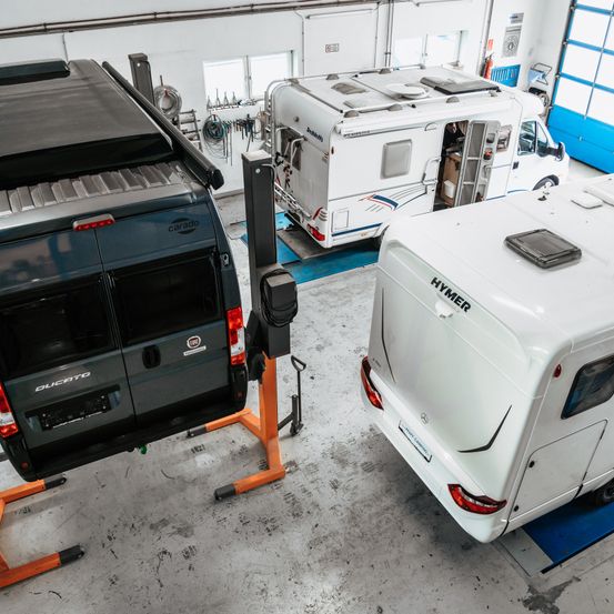 Eine Werkstatt mit drei Wohnmobilen. Ein Carado-Van steht auf einem Lift, ein Hymer-Wohnmobil steht auf dem Boden und ein weiteres weißes Wohnmobil ist geparkt.