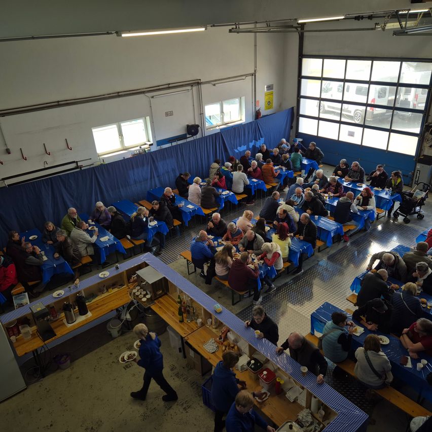 Eine Gruppe von Menschen diniert in einem Saal mit blau bedeckten Tischen. Einige Tische haben Essen und Getränke. Eine Frau serviert Essen.