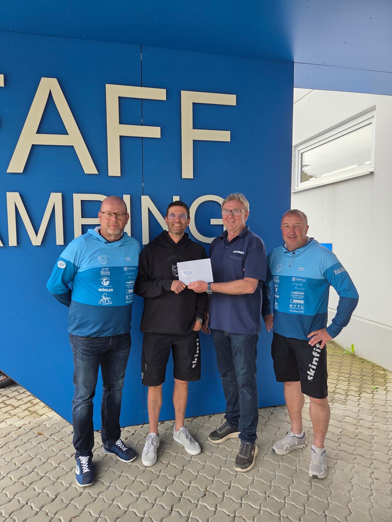 Vier Männer stehen vor einem blauen Schild mit dem Wort 'Personalcamping'. Drei tragen blaue Hemden, der in der Mitte trägt Schwarz. Sie halten ein Papier und posieren für ein Foto.