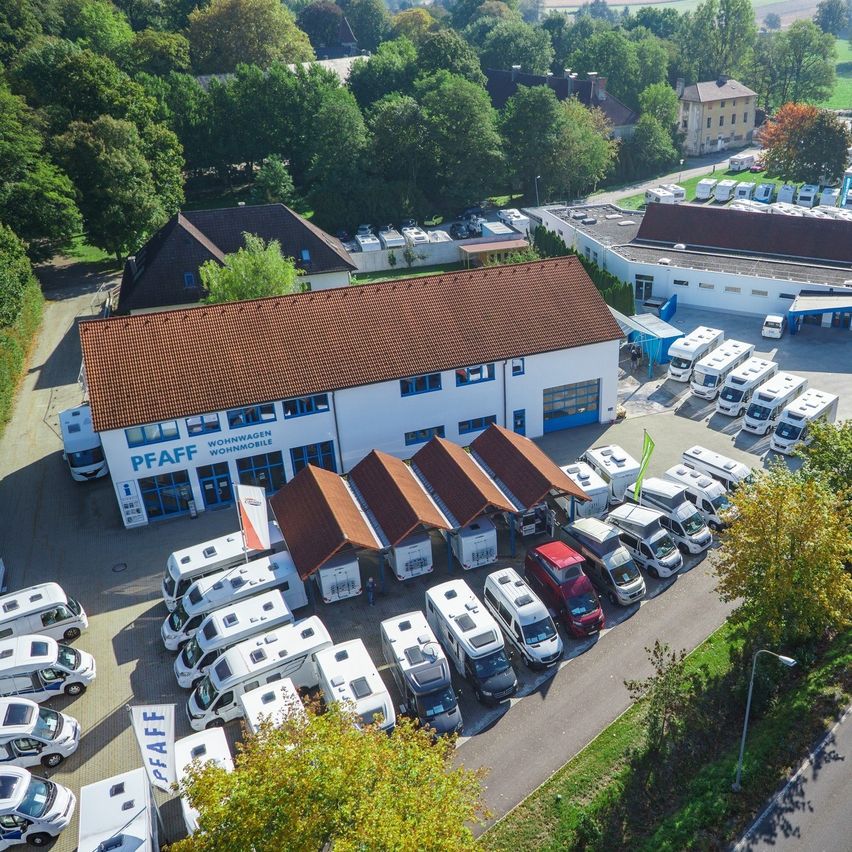 Luftaufnahme eines weissen Gebäudes mit braunen Dächern, beschriftet mit 'PFAFF', umgeben von zahlreichen geparkten Wohnmobilen und Autos, inmitten von Bäumen und Grün.