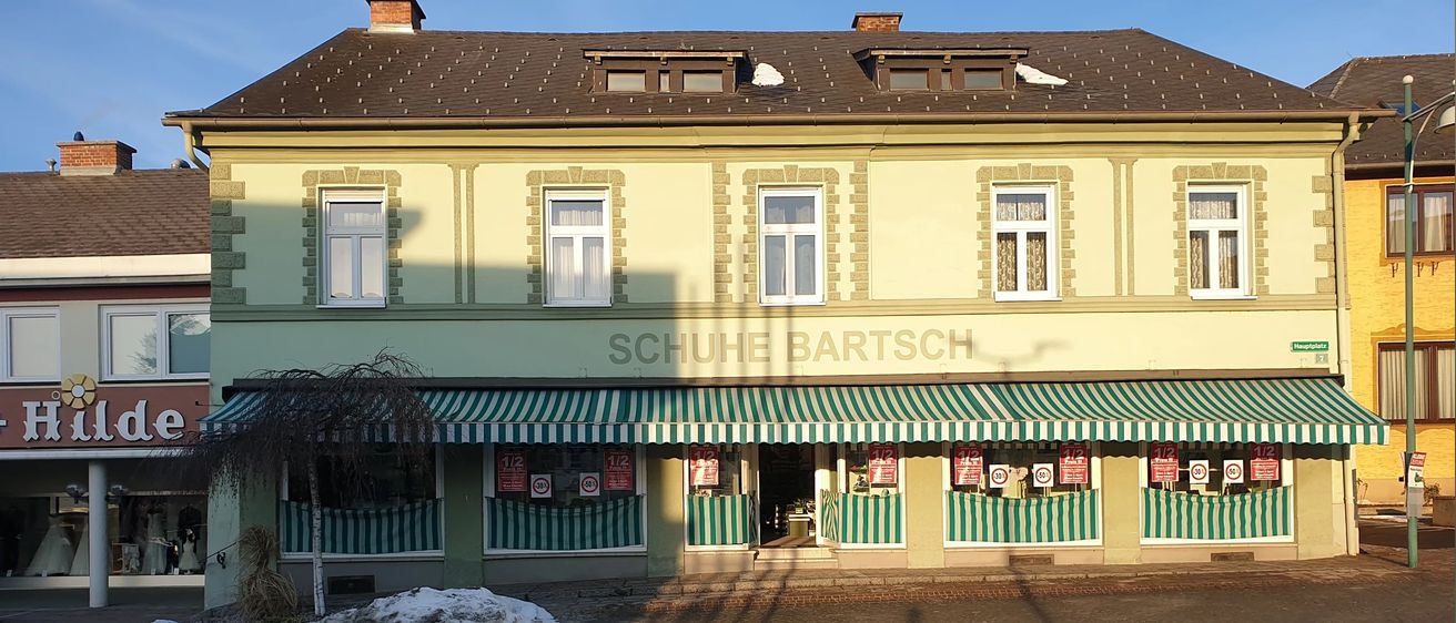 Ein Ladengeschäft mit einem grün-weiß gestreiften Vordach. Es hat mehrere Fenster und ist mit Schuh-Bartsch beschriftet. Vor dem Gebäude liegt Schnee auf dem Boden.