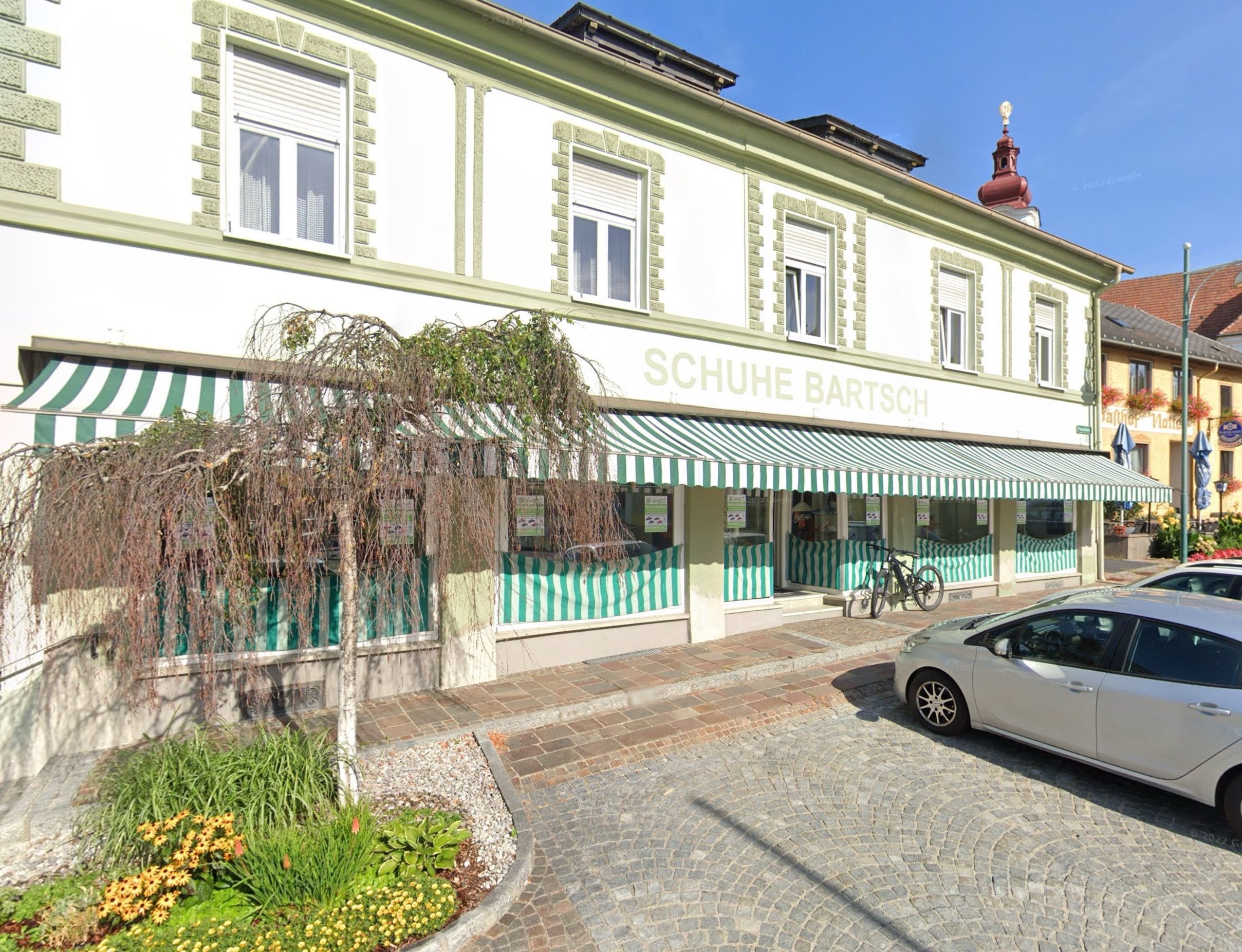 Ein weißes Gebäude mit grünem Vordach heißt Schuhie Bartsch. Es hat mehrere Fenster, ein Fahrrad vor der Tür und ein Auto davor.