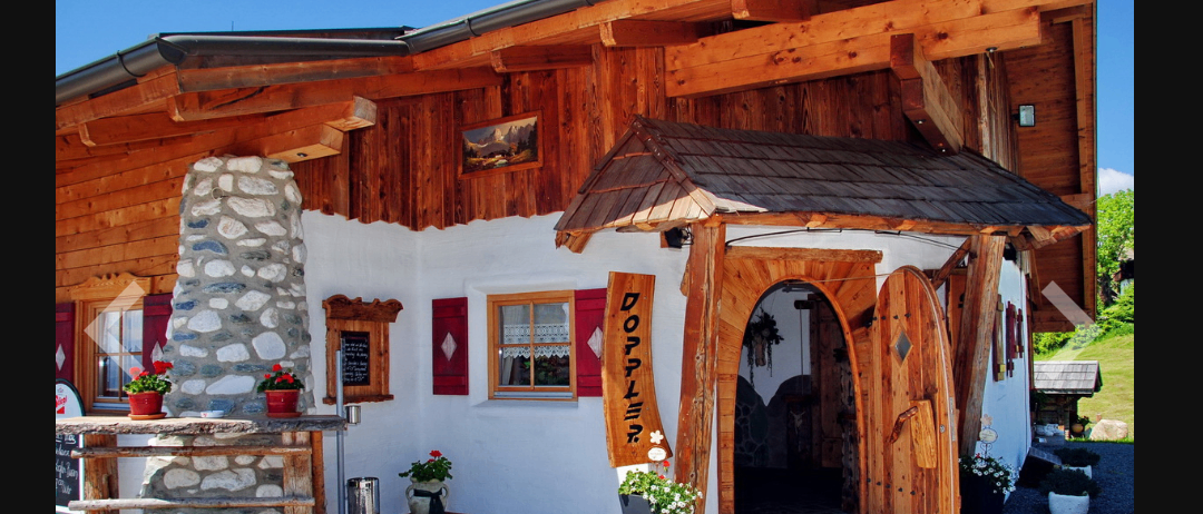 Ein Holzchalet mit einem Steinpfeiler und einer Holztür mit einem Schild, das DOPPLER steht. Draußen gibt es Topfpflanzen und Blumen.