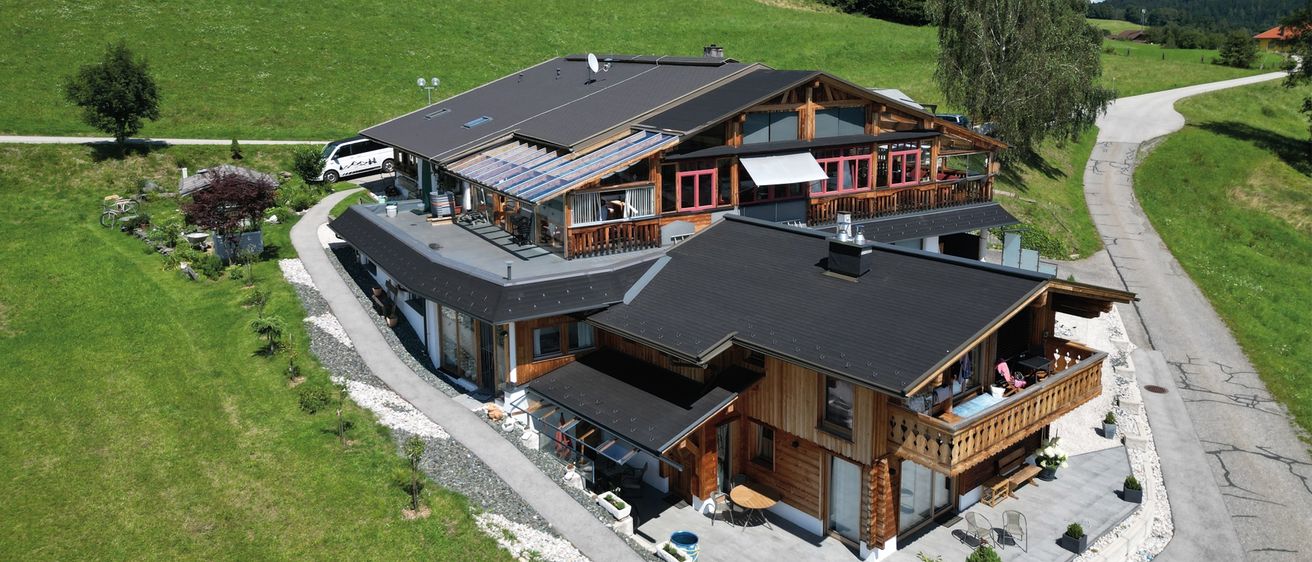 Ein Luftbild eines Chalets mit schwarzem Dach und rotem Rand, mit großen Fenstern, einem Balkon und einem geparkten Auto in der Nähe. Das Haus hat Solarpanele und ist von einem grünen Feld umgeben.