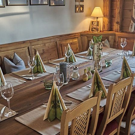 Ein hölzerner Esstisch ist mit Weingläsern, Servietten und einer Lampe besetzt. Holzstühle sind um den Tisch herum angeordnet. Kissen sind auf der Bank. Ein hölzerner Schrank hat einen Bilderrahmen und eine Lampe oben.