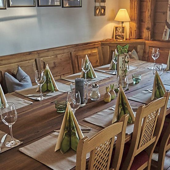 Ein hölzerner Esstisch ist mit Weingläsern, Servietten und einer Lampe besetzt. Holzstühle sind um den Tisch herum angeordnet. Kissen sind auf der Bank. Ein hölzerner Schrank hat einen Bilderrahmen und eine Lampe oben.