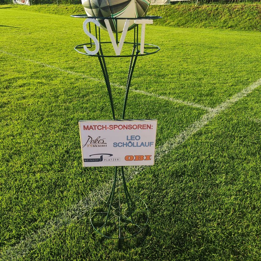 Ein Fußball auf einem Ständer mit dem Text 'SVT' in weiß. Darunter steht ein Schild mit der Aufschrift 'Match-Sponsoren: Pokers Leo Schollauf Weinhof Platzer'. Das Fußballfeld ist grün.