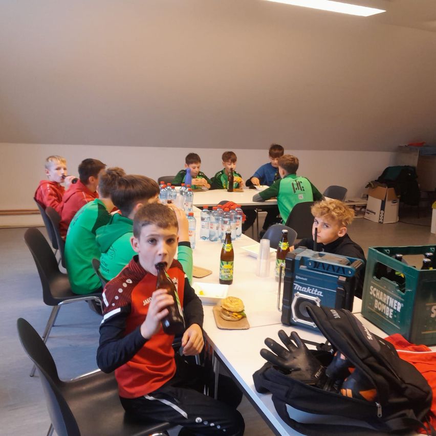Eine Gruppe von Jungen in grünen und roten Trikots sitzt an einem langen Tisch, einige trinken aus Flaschen. Ein Junge trinkt aus einer braunen Flasche. Ein Hamburger, Wasserflaschen und ein Makita-Lautsprecher stehen auf dem Tisch.