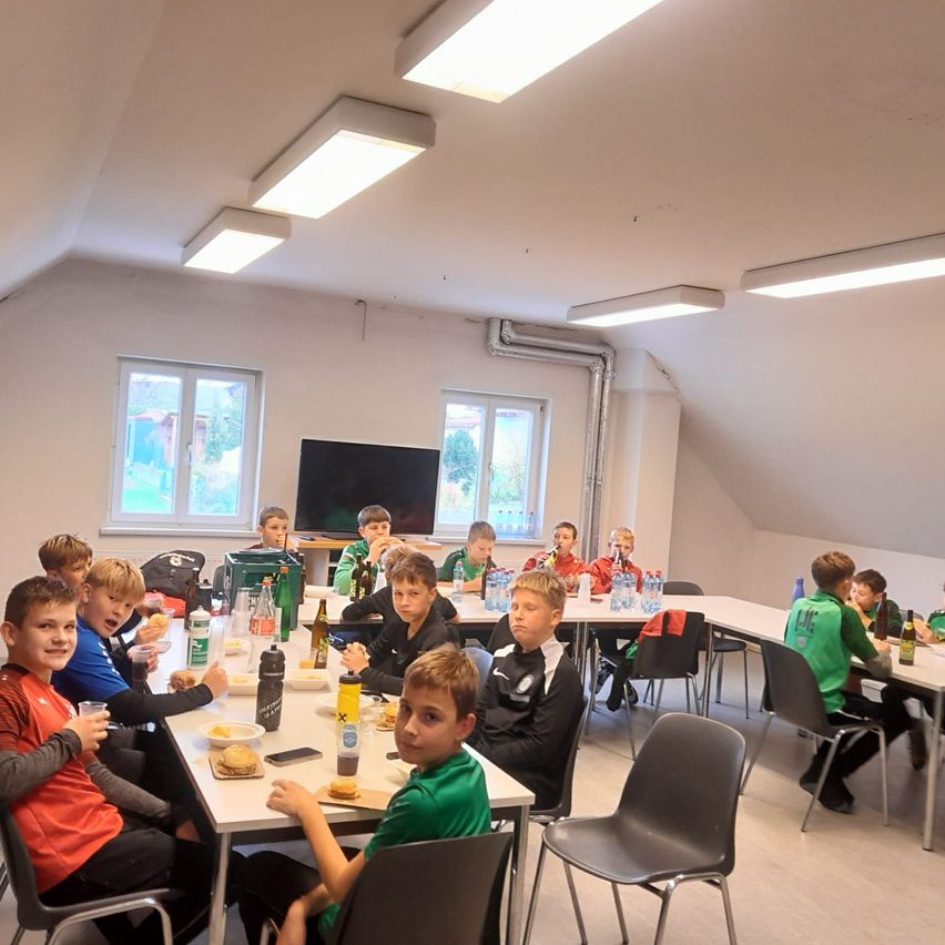 Eine Gruppe von Kindern sitzt an Tischen in einem Raum, einige essen und andere trinken. Der Raum hat weiße Wände und Decke mit Lampen, Fenster und einen Fernseher. Flaschen und Becher stehen auf den Tischen.