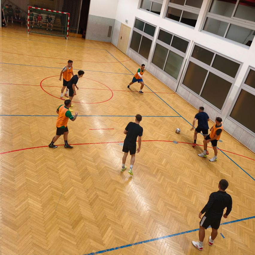 Eine Gruppe von Männern in Sportuniformen spielt Fußball auf einem Hallenplatz mit Holzböden. Der Platz hat rote und blaue Linien und ein Tor auf der linken Seite.