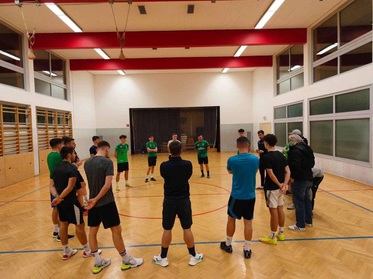 Eine Gruppe von Männern in grünen und schwarzen Uniformen steht in einem Kreis auf einem Innenspielfeld. Sie unterhalten sich wahrscheinlich miteinander. Das Spielfeld hat einen Holzboden und rote Markierungen. In der Ferne stehen auch Menschen. Die Decke hat rote Balken und weiße Lampen.