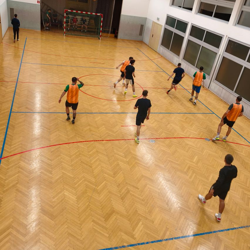 Eine Gruppe von Menschen in Sportuniformen spielt Futsal auf einem Innenplatz mit blauen und roten Markierungen. Zwei Personen sind in der Nähe des Tores.