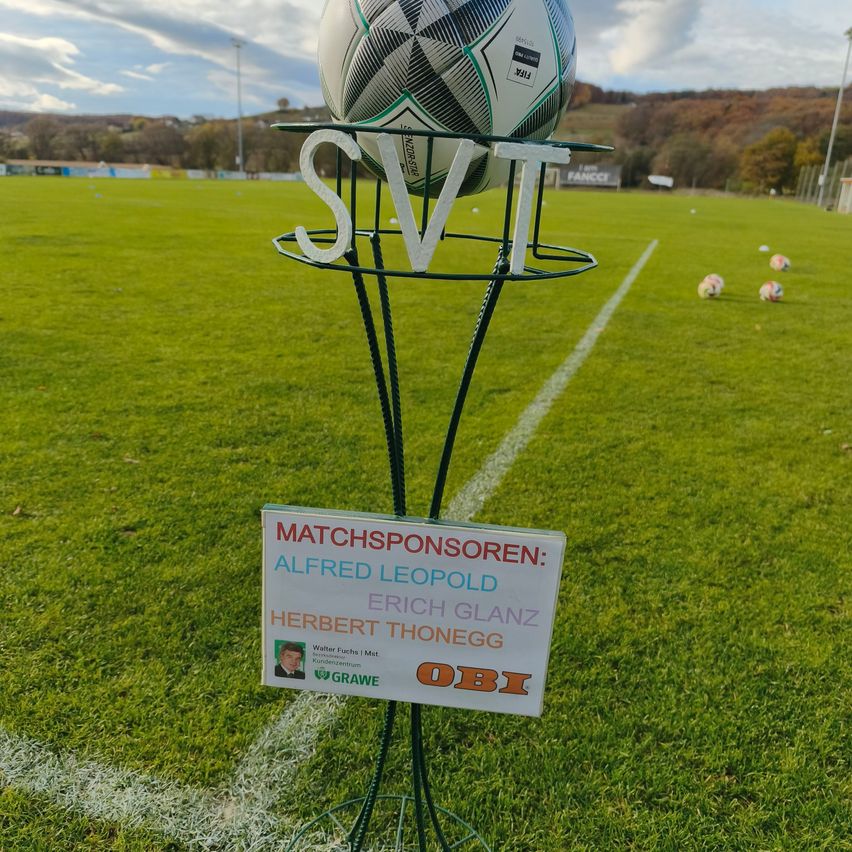 Ein Fußball steht auf einem Ständer auf einem Fußballfeld mit den Buchstaben SVT darüber. Es gibt ein Schild mit Namen.