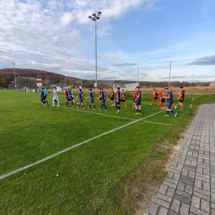 Ein Fußballspiel findet auf einem gut gepflegten Feld statt, mit Spielern in blauen und orangefarbenen Trikots. Das Feld hat eine weiße Linie und einen Ziegelweg auf der rechten Seite. Straßenlaternen und ein Netzzaun sind vorhanden.