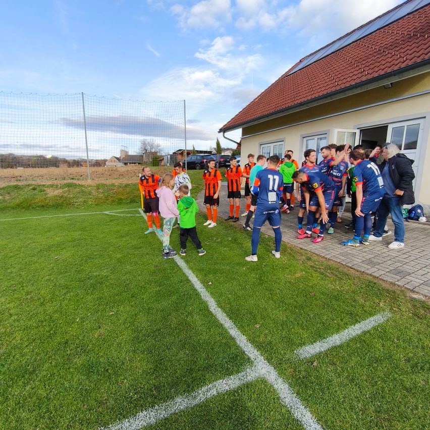 Eine Fußballmannschaft steht in der Nähe eines Hauses mit Solardach. Spieler in orangen und blauen Trikots versammeln sich auf dem Rasen.