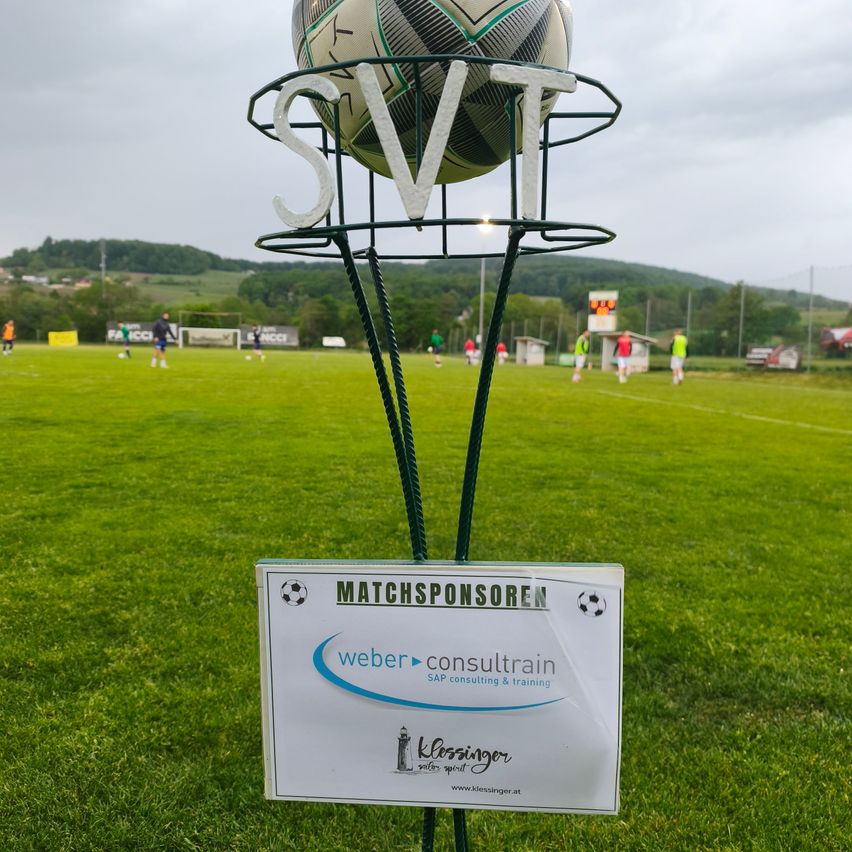 Ein Fußball wird von einem Ständer gehalten, der mit 'Matchsponseren' beschriftet ist. Im Hintergrund ist ein Fußballfeld mit Spielern zu sehen.