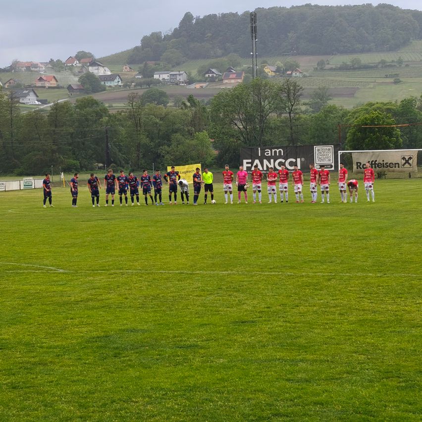 Fußballspieler in roten und weißen Trikots stehen auf dem Feld, möglicherweise für ein Foto posierend. Das Feld ist grün, und es gibt Bäume und Häuser im Hintergrund. Ein Schild steht für 'Ich bin FANCCI'.