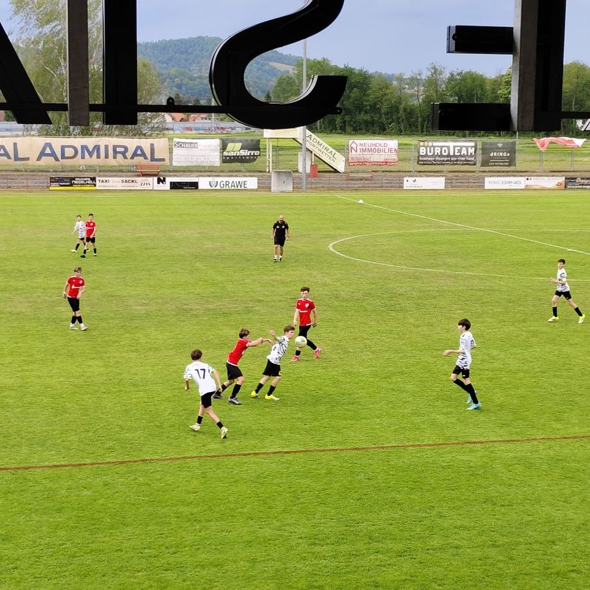 Ein Fußballspiel auf einem Feld mit Spielern in roten und weißen Trikots, einem Schiedsrichter und Zuschauern. Das Feld ist von Bannern und einem Zaun umgeben.